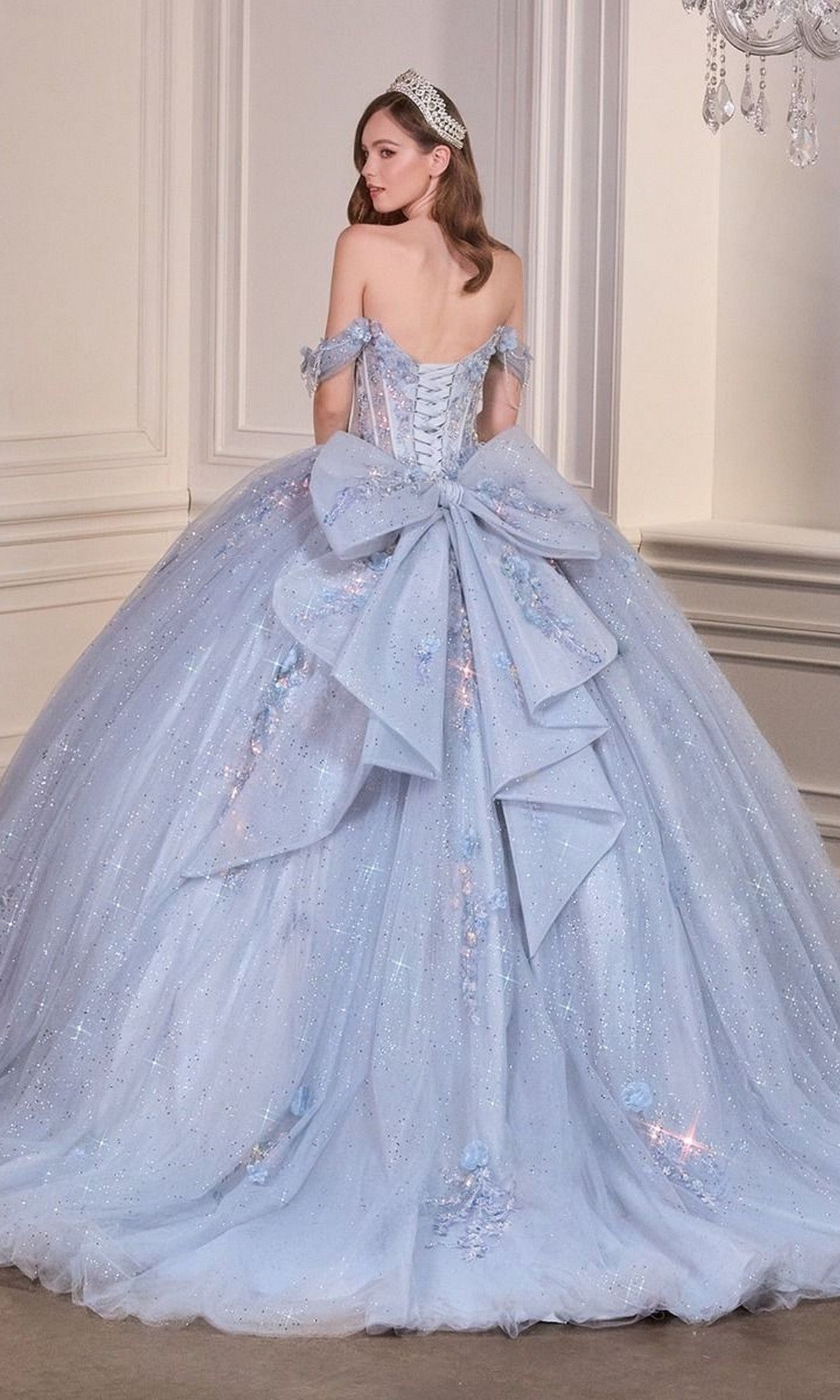 A woman wears the Quinceanera Dress 15728 By Ladivine, a light blue off-the-shoulder ball gown with sparkling details and a large back bow, standing in an elegant room.